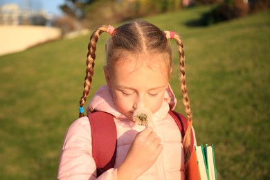 Photo of Cute little girl with dandelion, backpack and textbooks outdoors