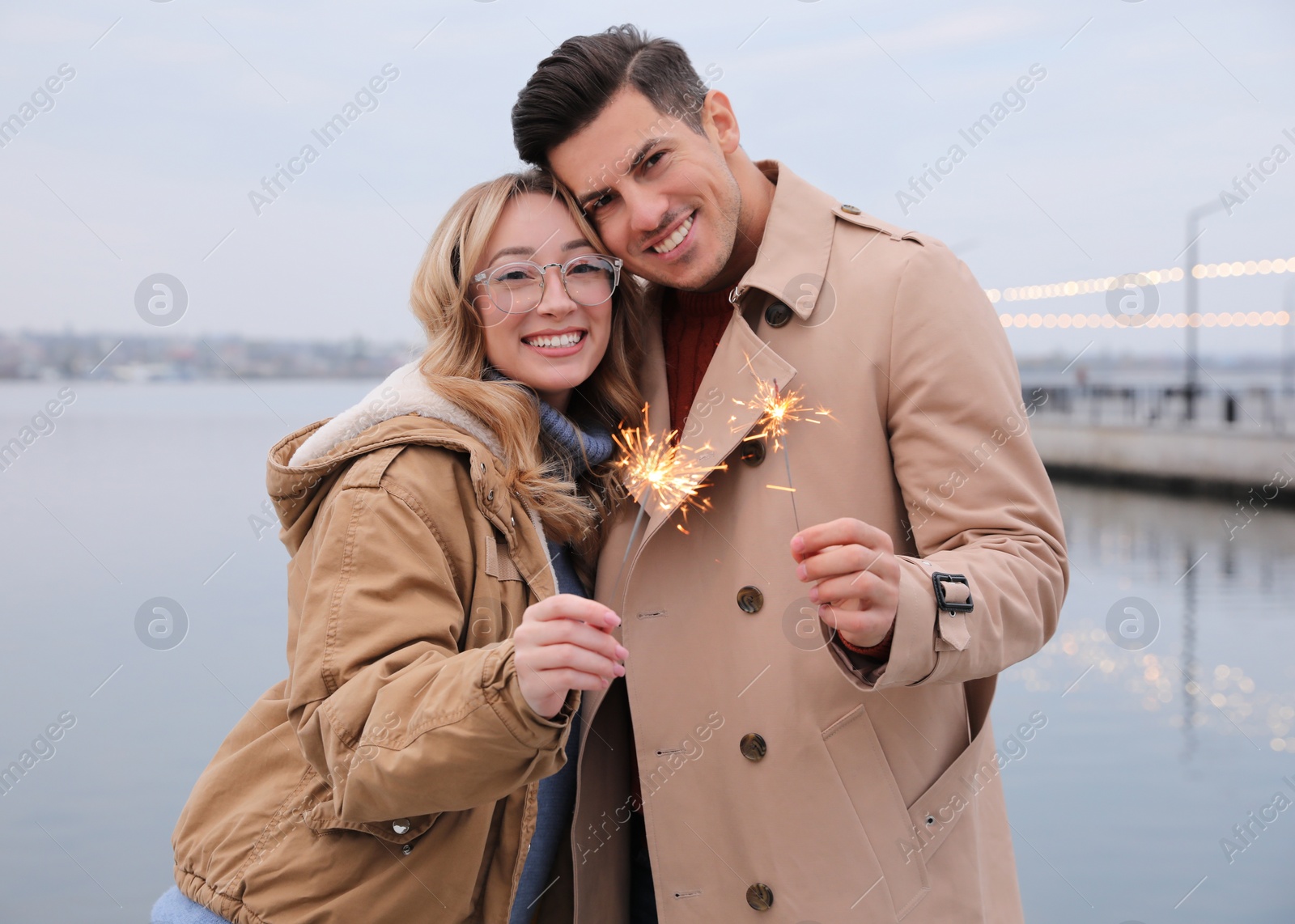 Photo of Couple in warm clothes holding burning sparklers near river