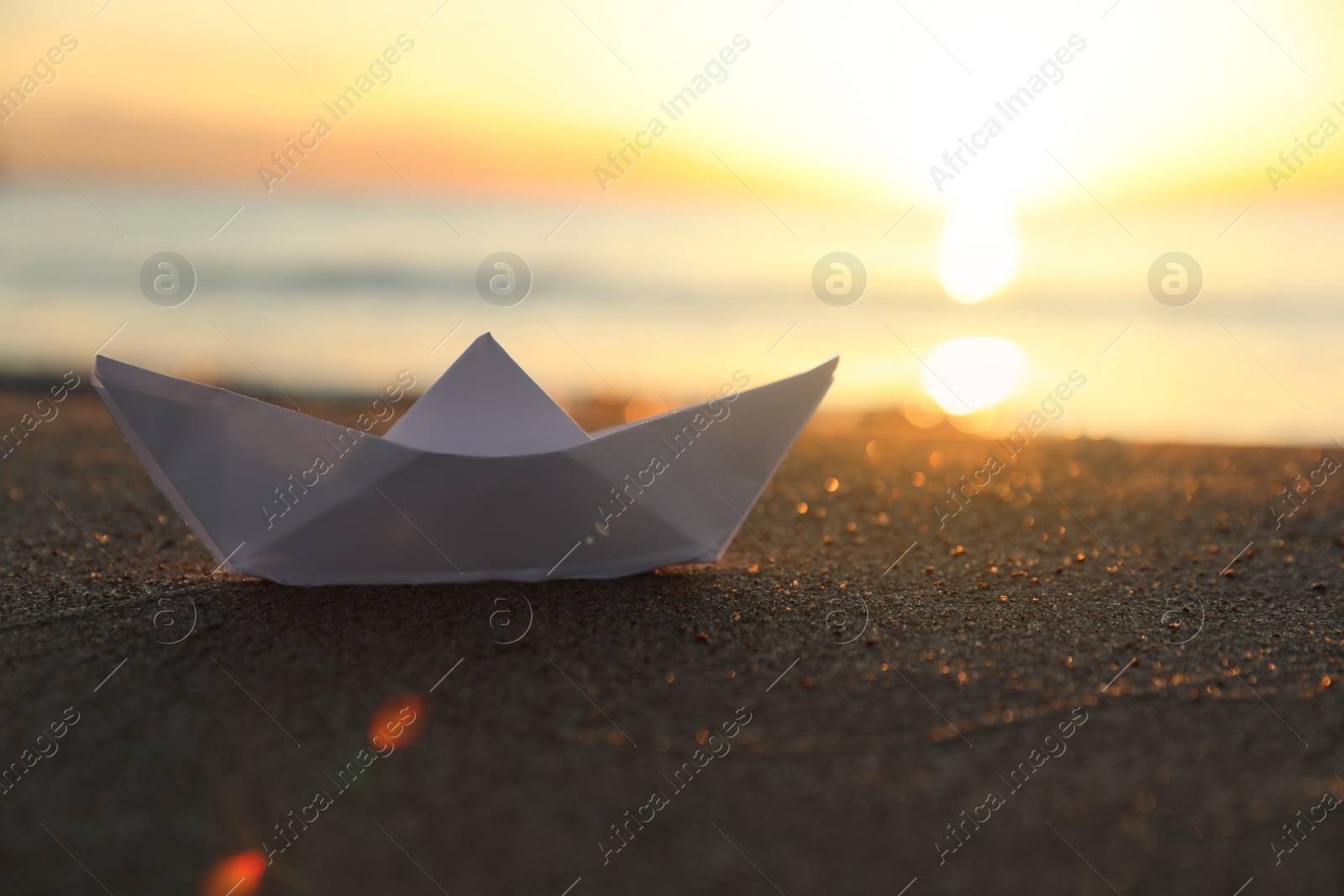 Photo of White paper boat on sand near sea at sunset, closeup. Space for text