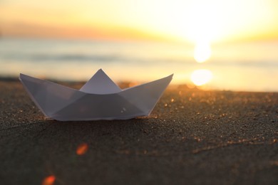 Photo of White paper boat on sand near sea at sunset, closeup. Space for text