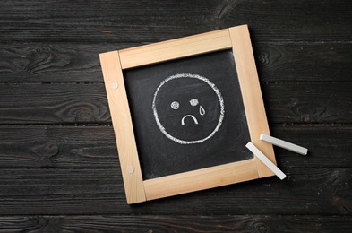 Photo of Small chalkboard with drawing of sad crying face on wooden background, top view. Depression symptoms