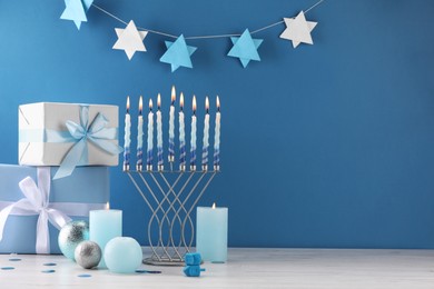 Photo of Hanukkah celebration. Menorah, burning candles, dreidels and gift boxes on white wooden table against light blue background, space for text