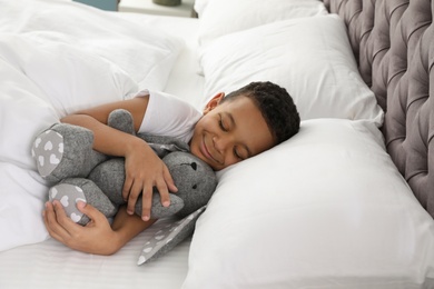 Cute little African-American boy with toy rabbit sleeping in bed