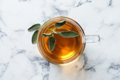 Cup of aromatic sage tea with fresh leaves on white marble table, top view