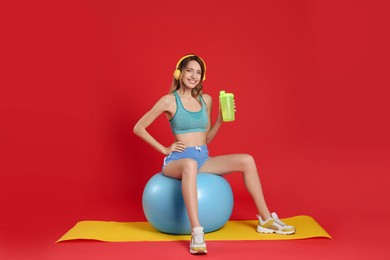 Photo of Beautiful woman with headphones and shaker sitting on fitness ball against red background