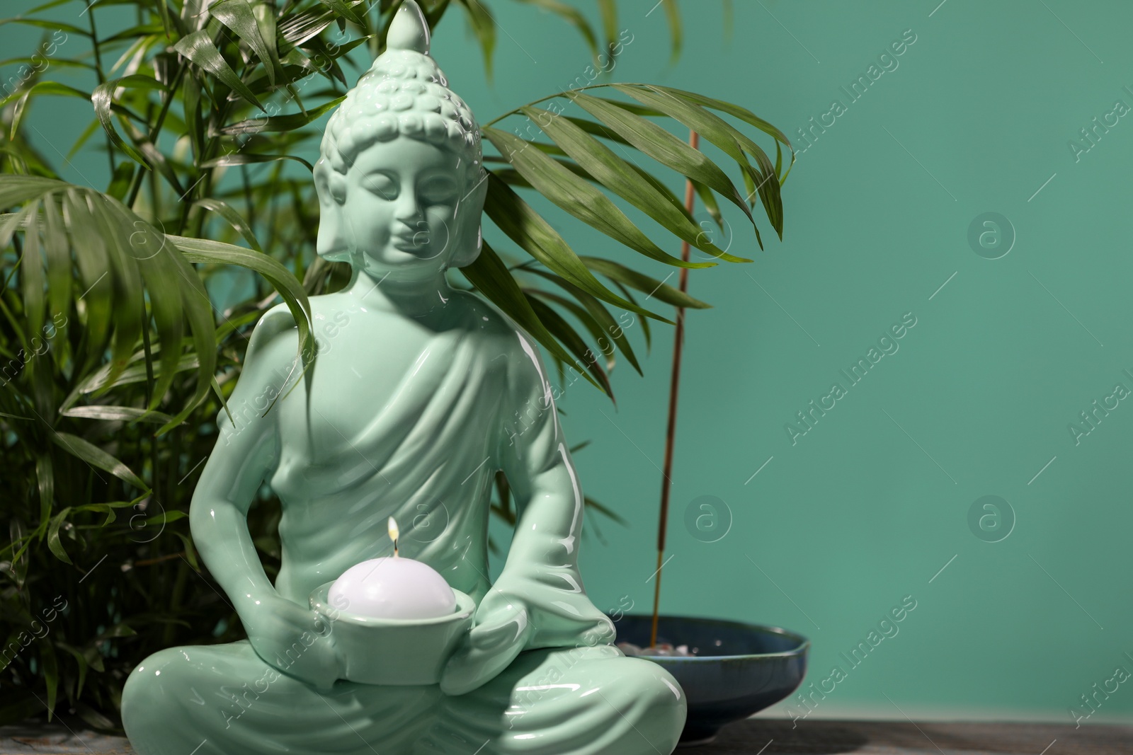 Photo of Buddhism religion. Decorative Buddha statue with burning candle on table and houseplant against turquoise wall, space for text