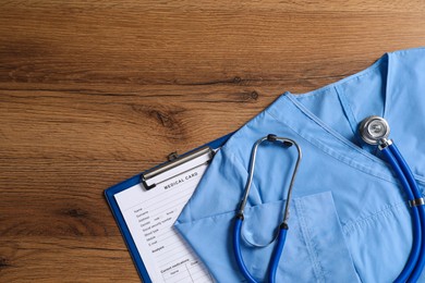 Photo of Medical uniform, stethoscope and clipboard on wooden table, flat lay. Space for text