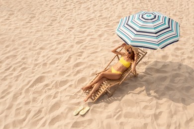 Woman resting in sunbed under striped beach umbrella at sandy coast, space for text