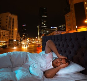 Annoyed man in bed and beautiful view of night cityscape on background. Poor sleep because of urban noise