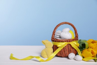 Photo of Wicker basket with festively decorated Easter eggs, bunny and beautiful tulips on white wooden table against light blue background. Space for text