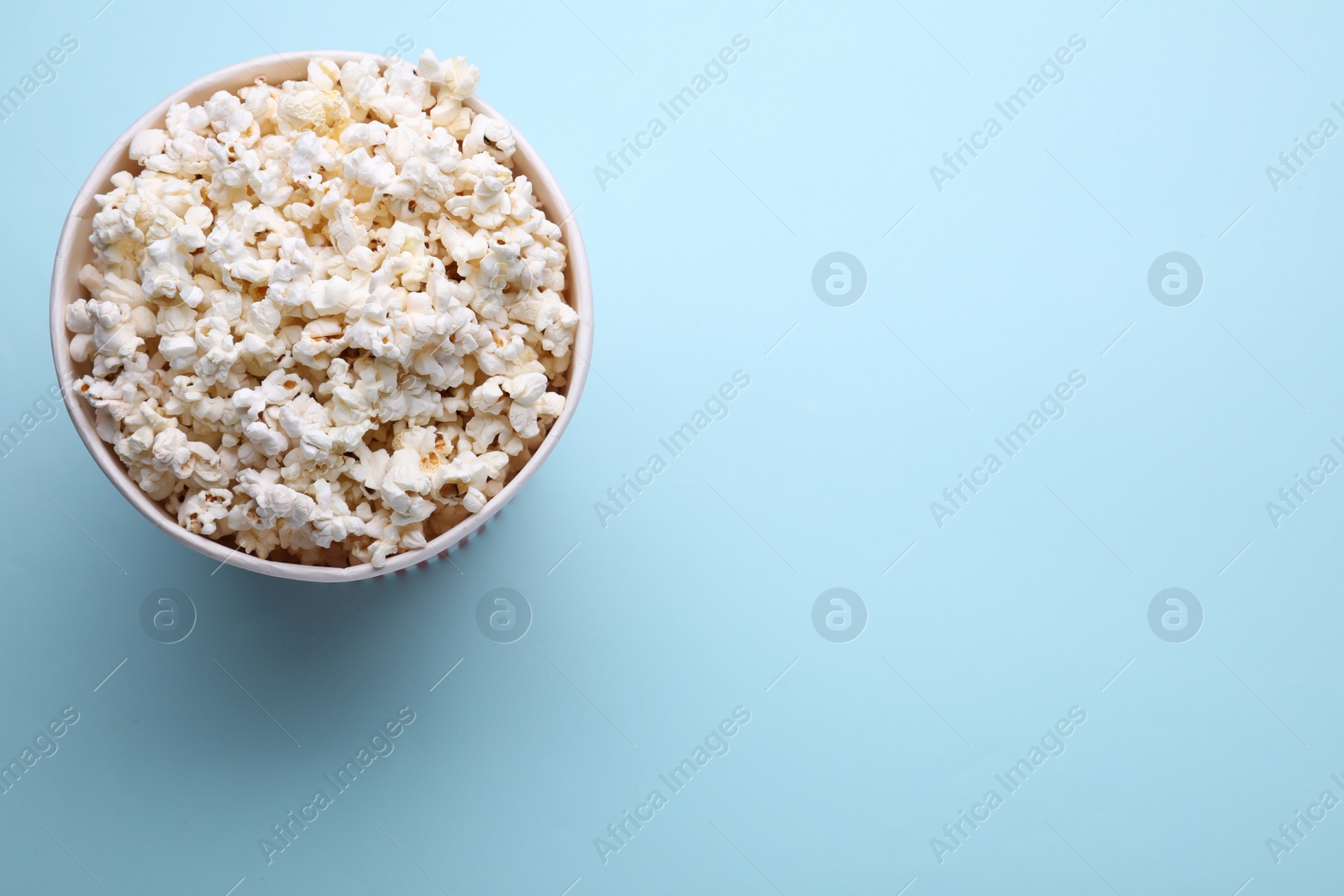 Photo of Paper bucket with delicious popcorn on light blue background, top view. Space for text