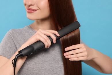 Woman using hair iron on light blue background, closeup