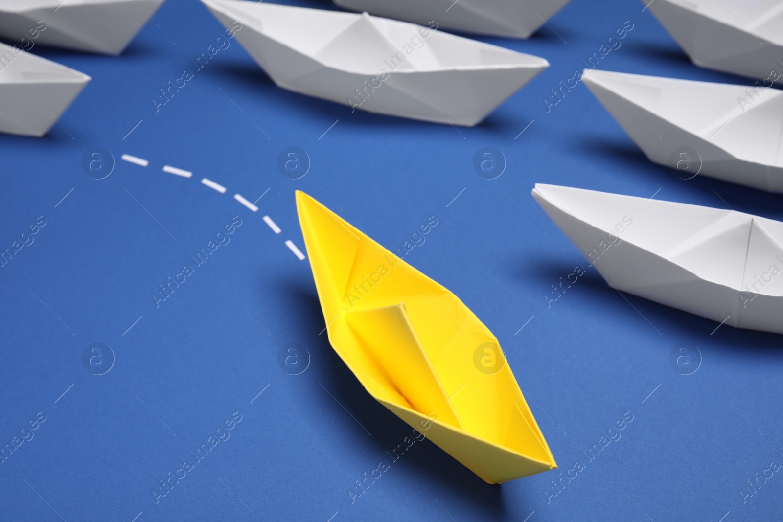 Photo of Yellow paper boat floating away from others on blue background, closeup. Uniqueness concept