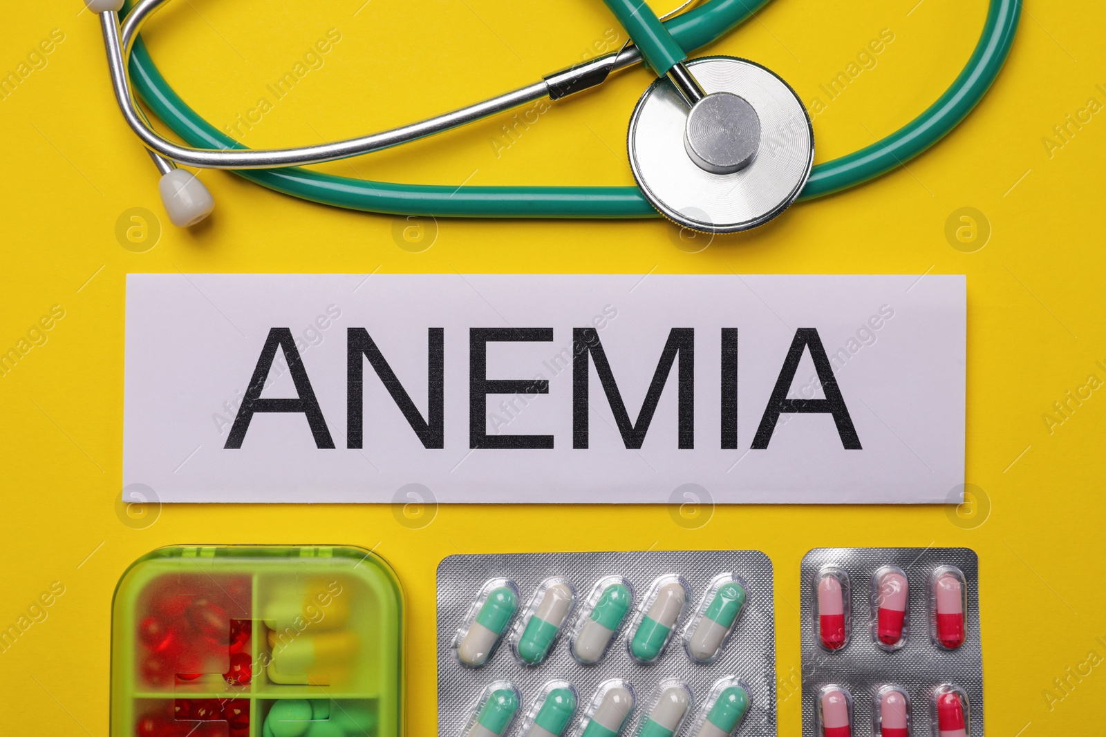 Photo of Card with word Anemia, different pills and stethoscope on yellow background, flat lay