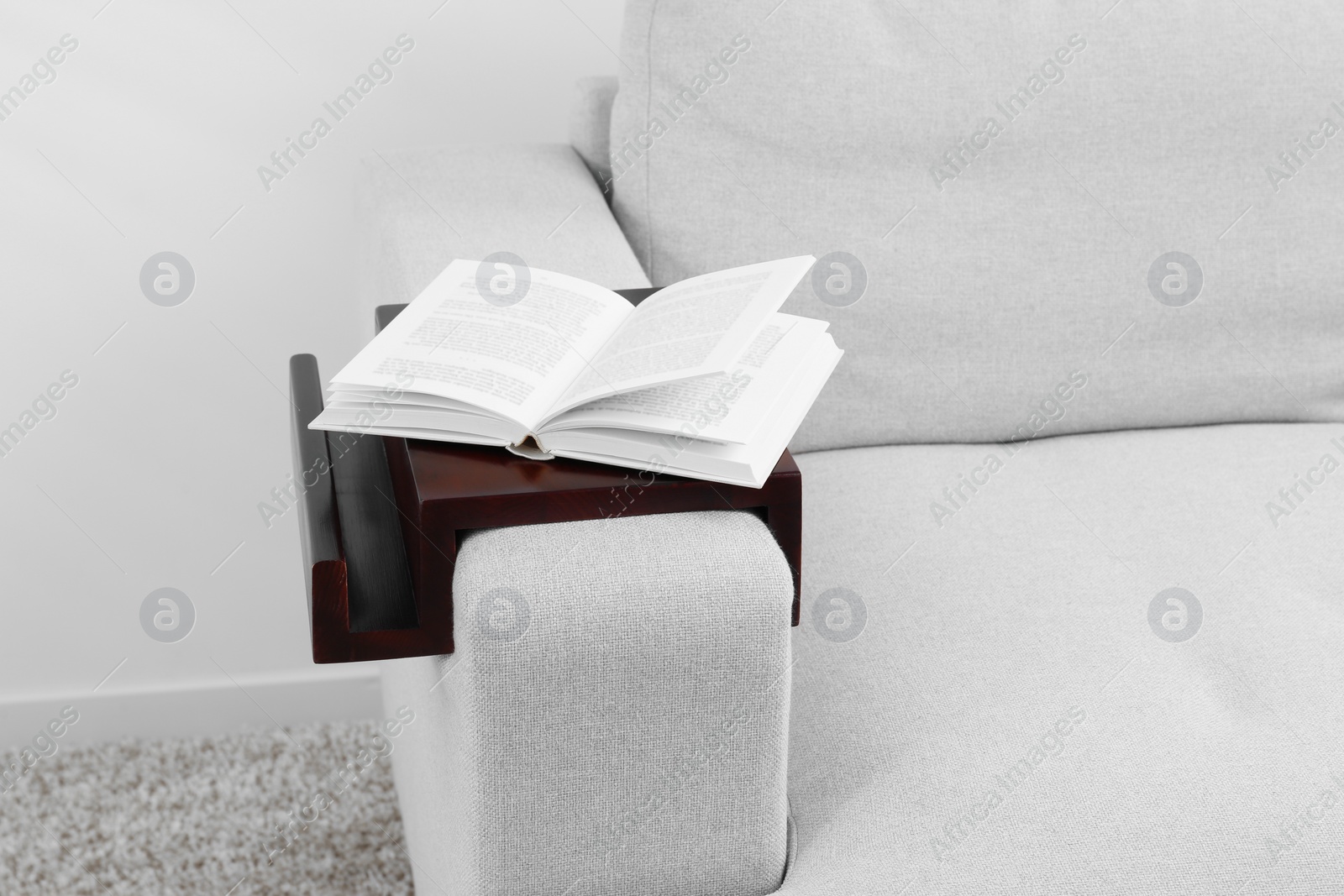 Photo of Open book on sofa with wooden armrest table indoors. Interior element