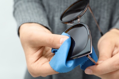 Woman wiping sunglasses with microfiber cleaning cloth on light background, closeup