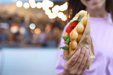 Photo of Young woman holding delicious bubble waffle with tomato and arugula outdoors, closeup