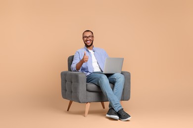 Photo of Smiling young man with laptop sitting in armchair and showing thumbs up on beige background