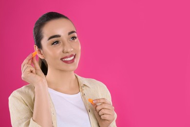 Young woman inserting foam ear plugs on pink background. Space for text