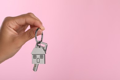 Photo of Woman holding key with metallic keychain in shape of house on pink background, closeup. Space for text