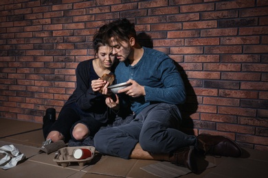 Photo of Poor young couple with bread on floor near brick wall