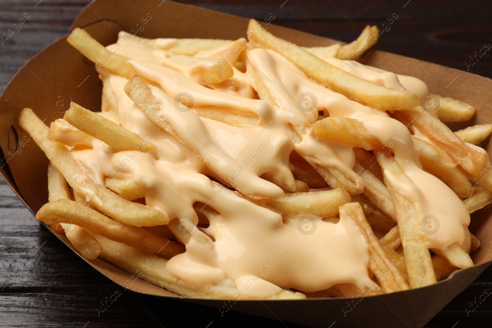 Photo of Tasty potato fries and cheese sauce in paper container on wooden table, closeup