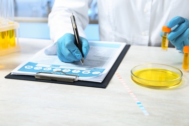 Photo of Laboratory assistant with urine sample for analysis writing results of tests at table indoors, closeup