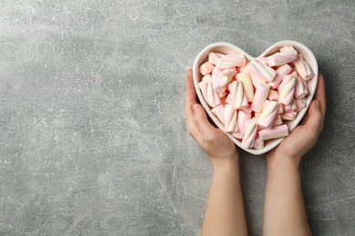 Photo of Woman holding heart shaped bowl full of marshmallows on grey background, top view. Space for text