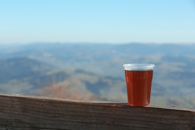 Photo of Plastic cup of natural hot tea on wooden railing in mountains. Space for text