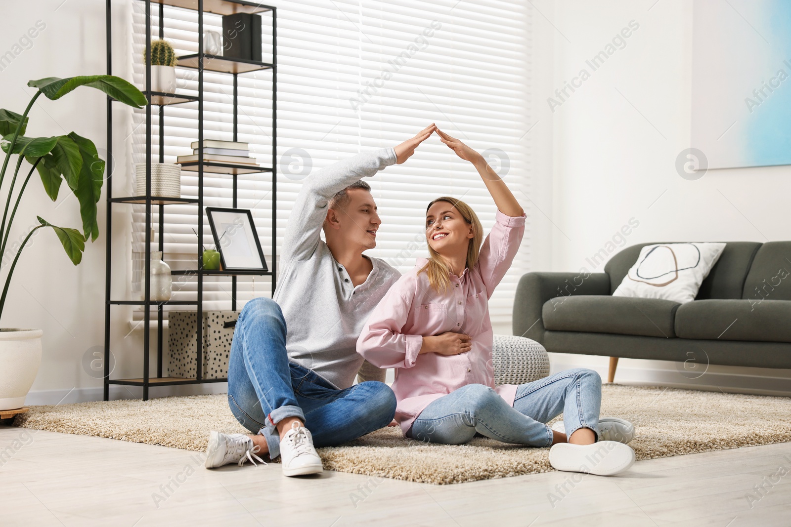 Photo of Young family housing concept. Pregnant woman with her husband forming roof with their hands while sitting on floor at home