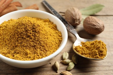 Photo of Dry curry powder in bowl, spoon and other spices on wooden table, closeup