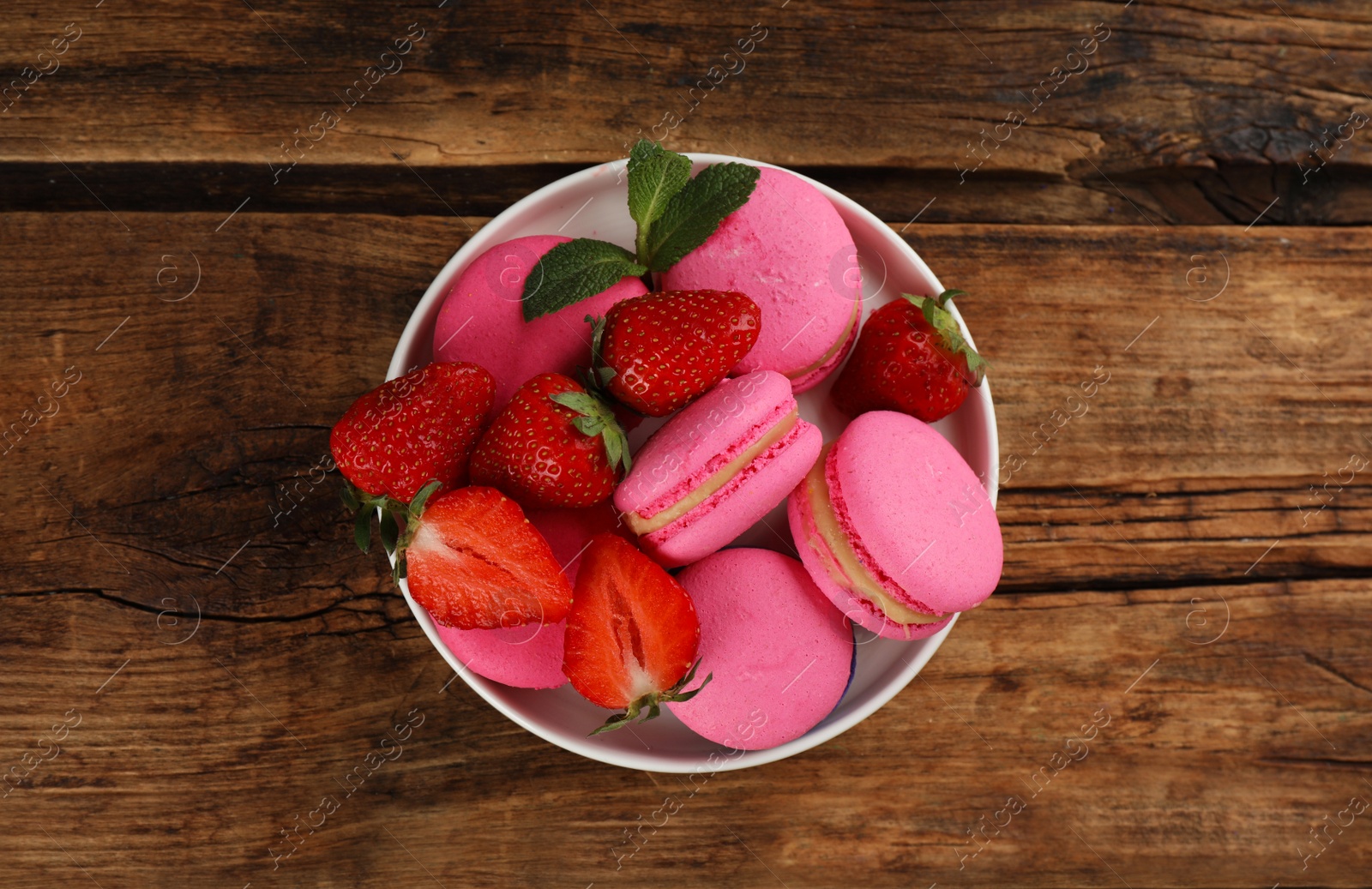 Photo of Delicious pink macarons, fresh strawberries and mint in bowl on wooden table, top view