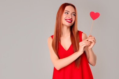 Photo of Young woman in red dress with paper heart on light grey background, space for text