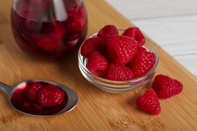 Photo of Jar of tasty canned raspberry jam and fresh berries in glass bowl on wooden table, closeup