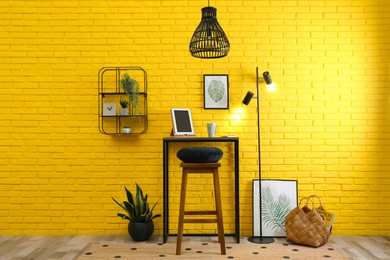 Photo of High wooden table with tablet and stool as stand up workplace near yellow brick wall. Stylish interior