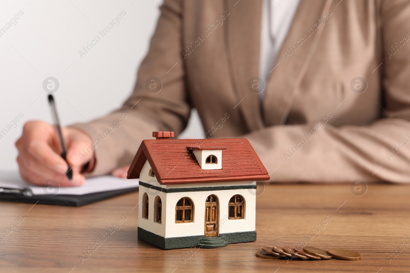 Photo of Mortgage concept. Woman writing something in notebook, house model and coins on wooden table, selective focus