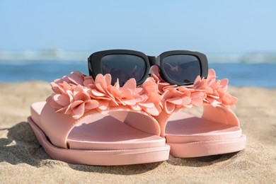 Stylish slippers and sunglasses on sandy beach near sea, closeup