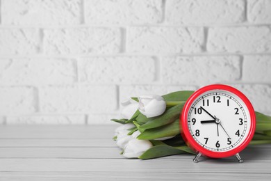 Photo of Red alarm clock and beautiful tulips on white wooden table against brick wall, space for text. Spring time