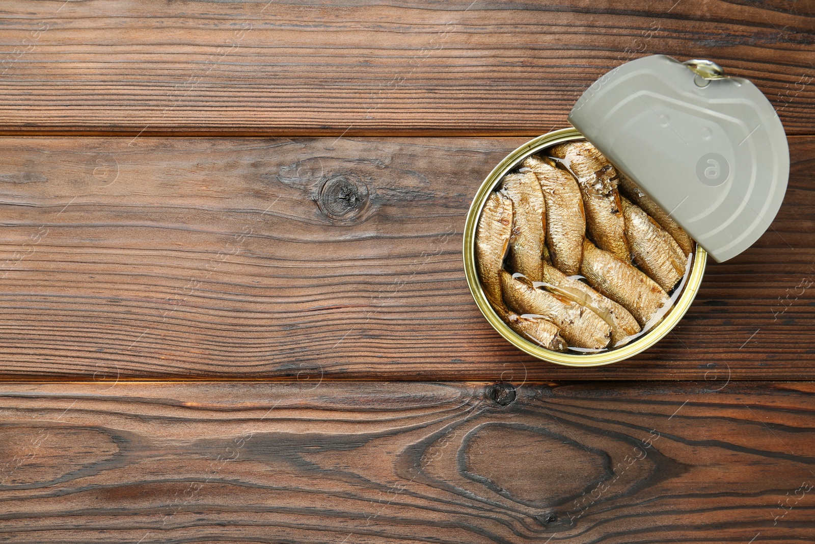 Photo of Open tin can of sprats on wooden table, top view. Space for text