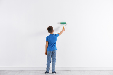 Photo of Little child painting with roller brush on white wall indoors