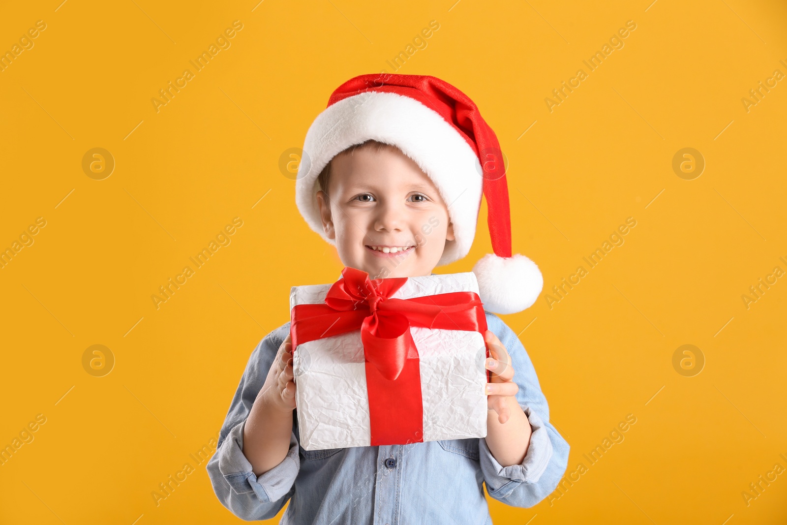 Photo of Cute little boy in Santa Claus hat holding gift box on yellow background