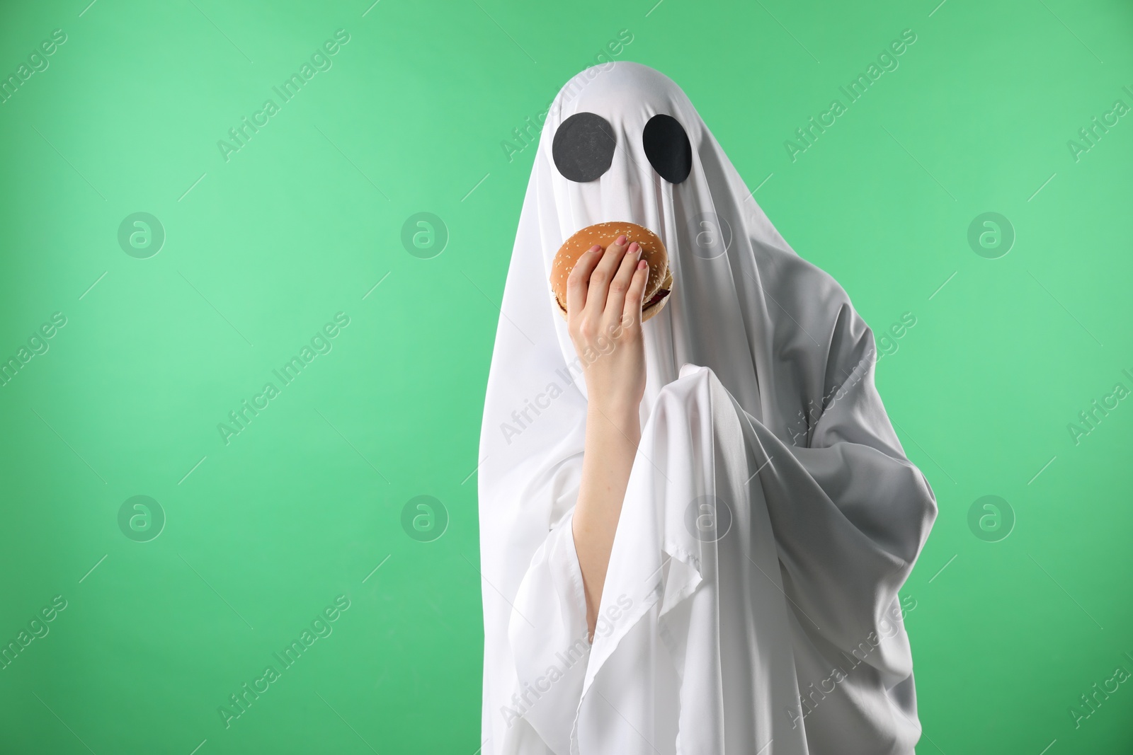 Photo of Creepy ghost. Person in white sheet with tasty hamburger on green background, space for text