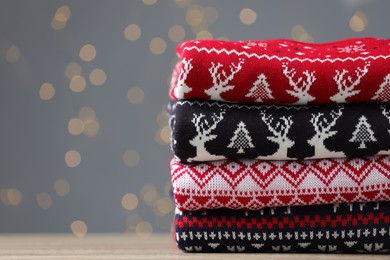 Stack of different Christmas sweaters on table against grey background with blurred lights. Space for text
