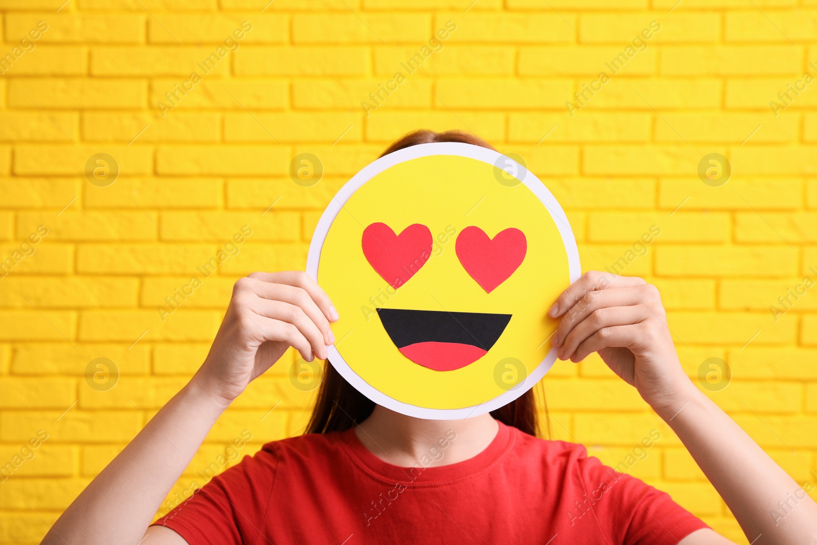 Photo of Woman covering face with heart eyes emoji near yellow brick wall