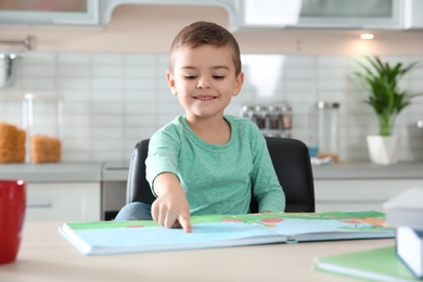 Cute little boy reading book at table in kitchen