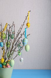 Photo of Beautiful willow branches with painted eggs in pot on light blue table, space for text. Easter decor