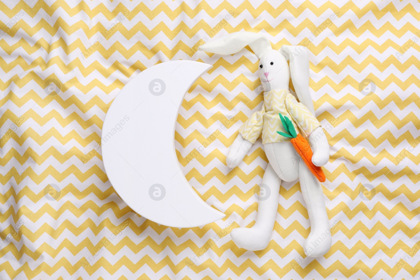 Photo of Crescent shaped child's night lamp and toy rabbit on yellow fabric, flat lay