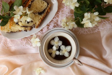 Photo of Cup of aromatic tea, tasty dessert and beautiful jasmine flowers on pink fabric, flat lay