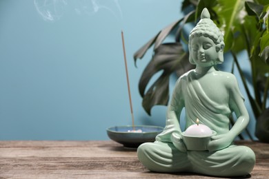 Buddhism religion. Decorative Buddha statue with burning candle on wooden table against light blue wall, space for text