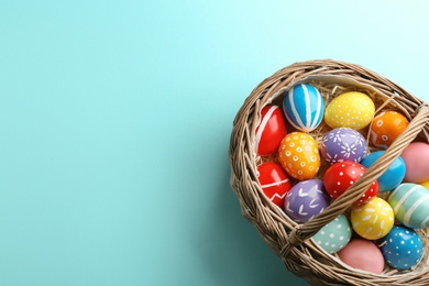 Wicker basket with painted Easter eggs on color background, top view. Space for text